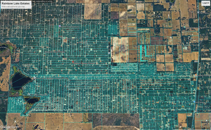 Aerial satellite view of a grid-like residential area labeled Rainbow Lake Estates, with many rectangular lots covered in bright green vegetation or fencing, surrounded by brown and tan farm fields.